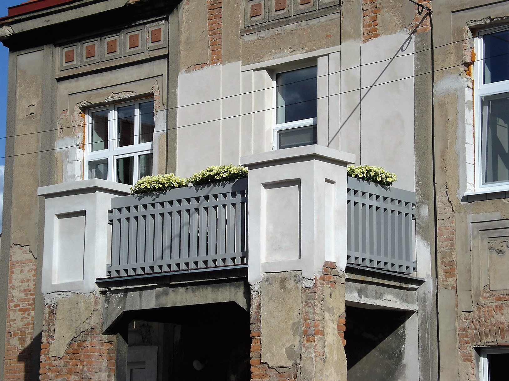 Celkový pohled zleva na balkon s šedým kovovým zábradlím navazujícím na zděné pilíře.