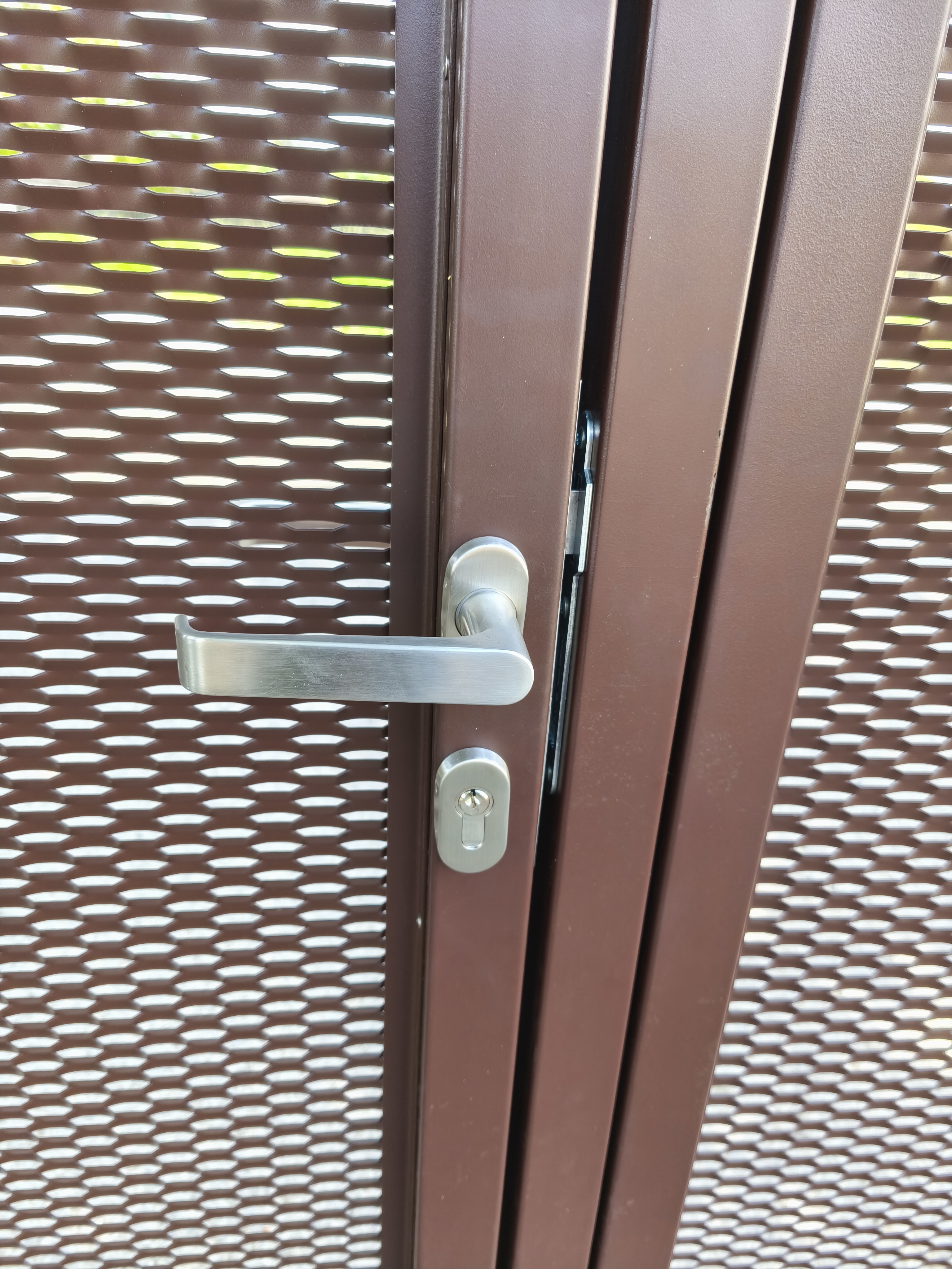 Detail kliky a cylindrické vložky branky integrované do hnědé tahokovové brány.