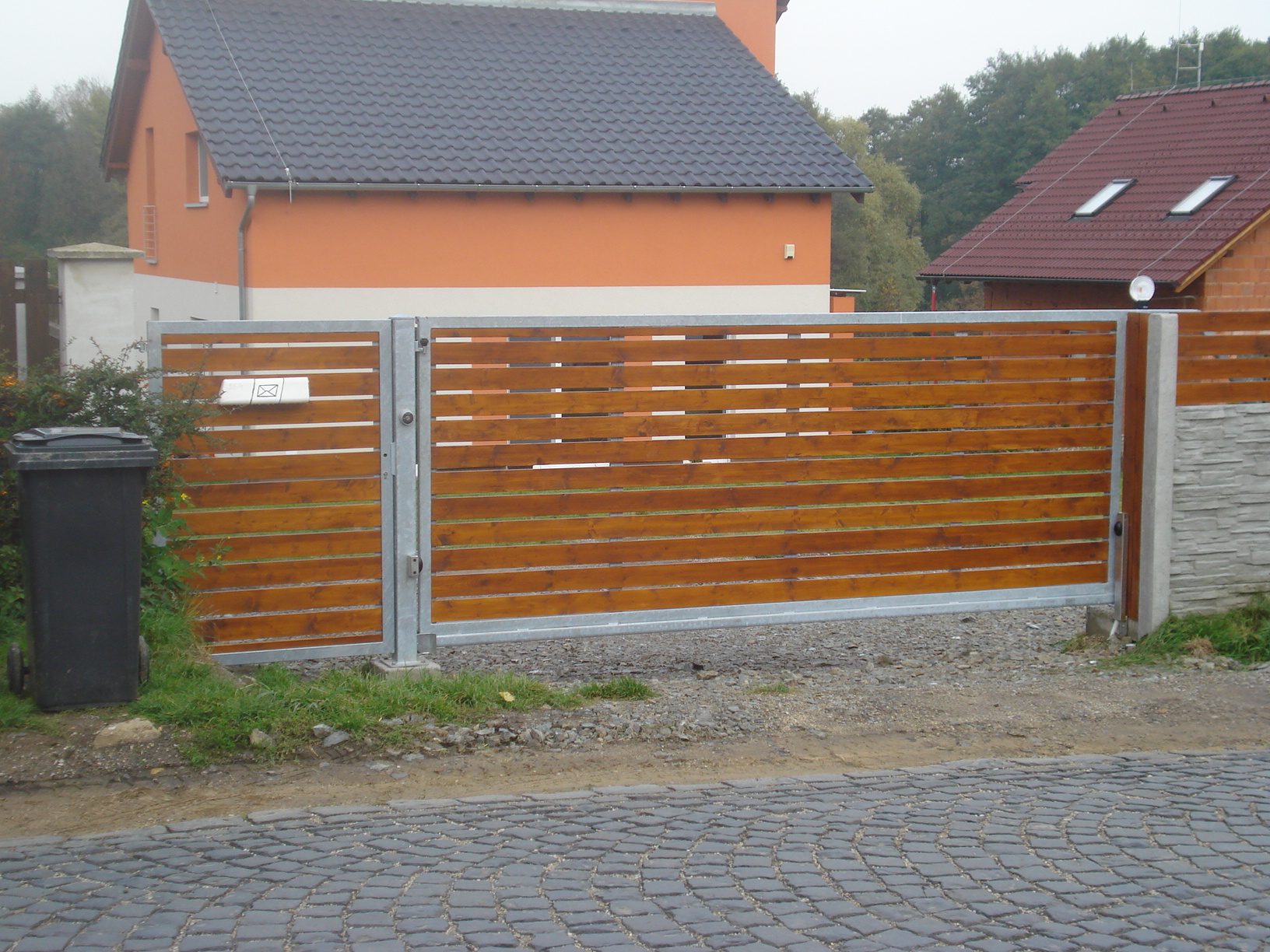 Celkový pohled z ulice na samonosnou bránu a branku s pozinkovanými rámy a vodorovnou dřevěnou výplní.