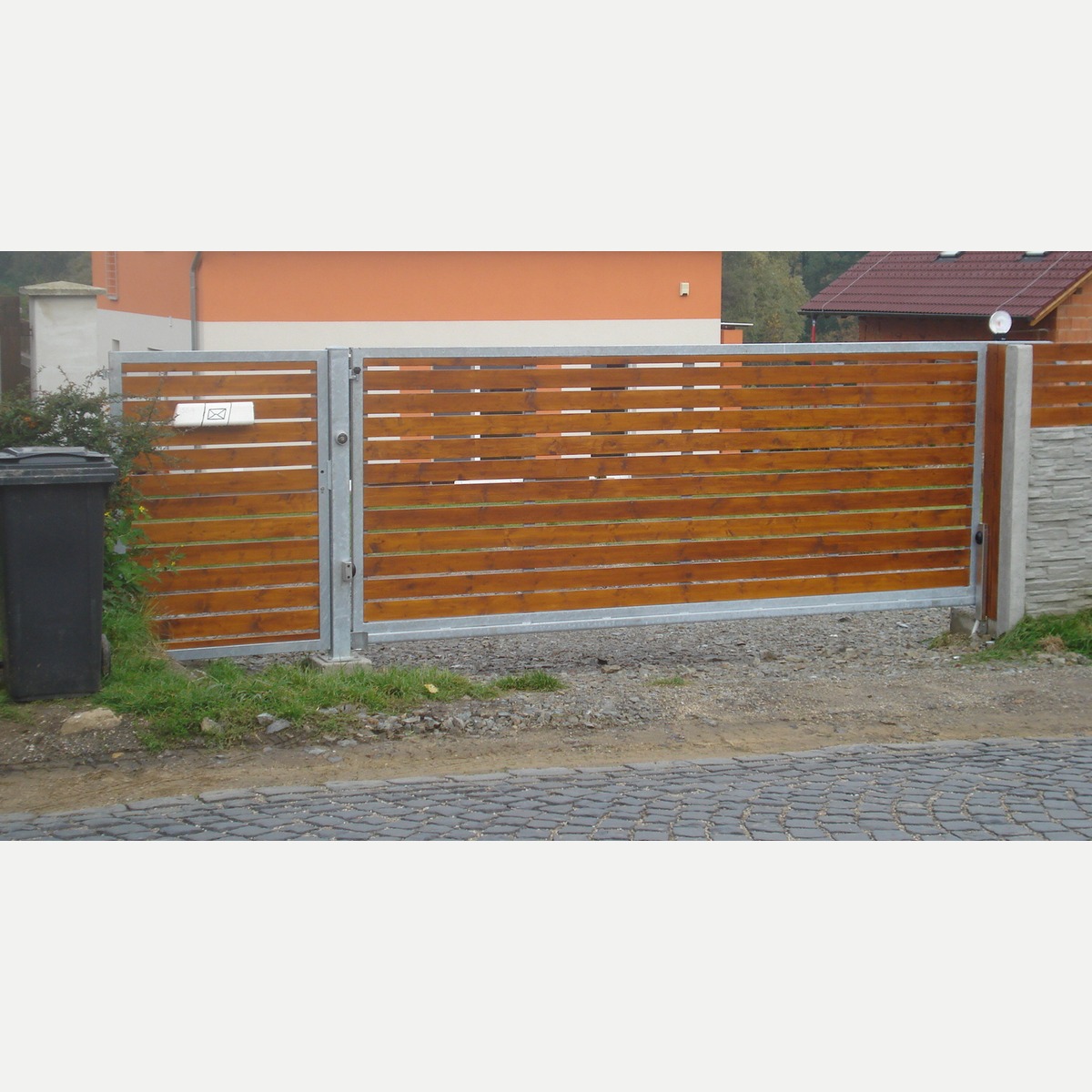 Celkový pohled z ulice na samonosnou bránu a branku s pozinkovanými rámy a vodorovnou dřevěnou výplní.