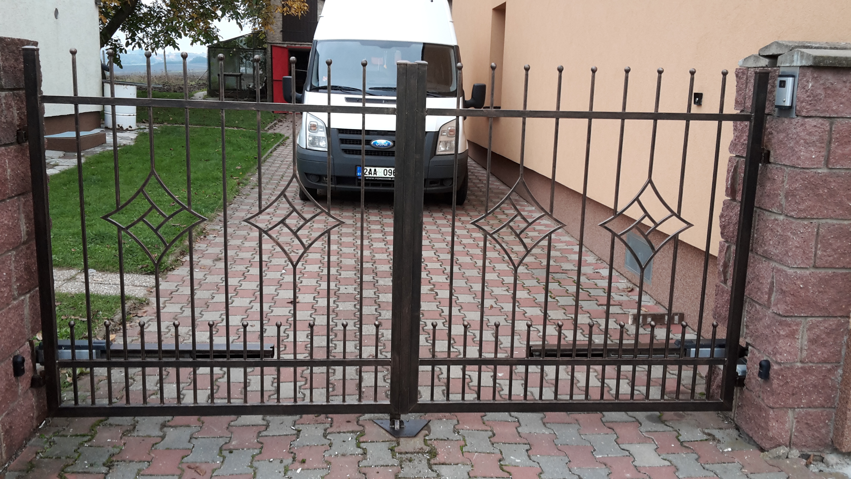 Čelní pohled na dvoukřídlou bránu v Hrdlívě se svislými pruty a geometrickými ozdobami mezi zděnými sloupky.