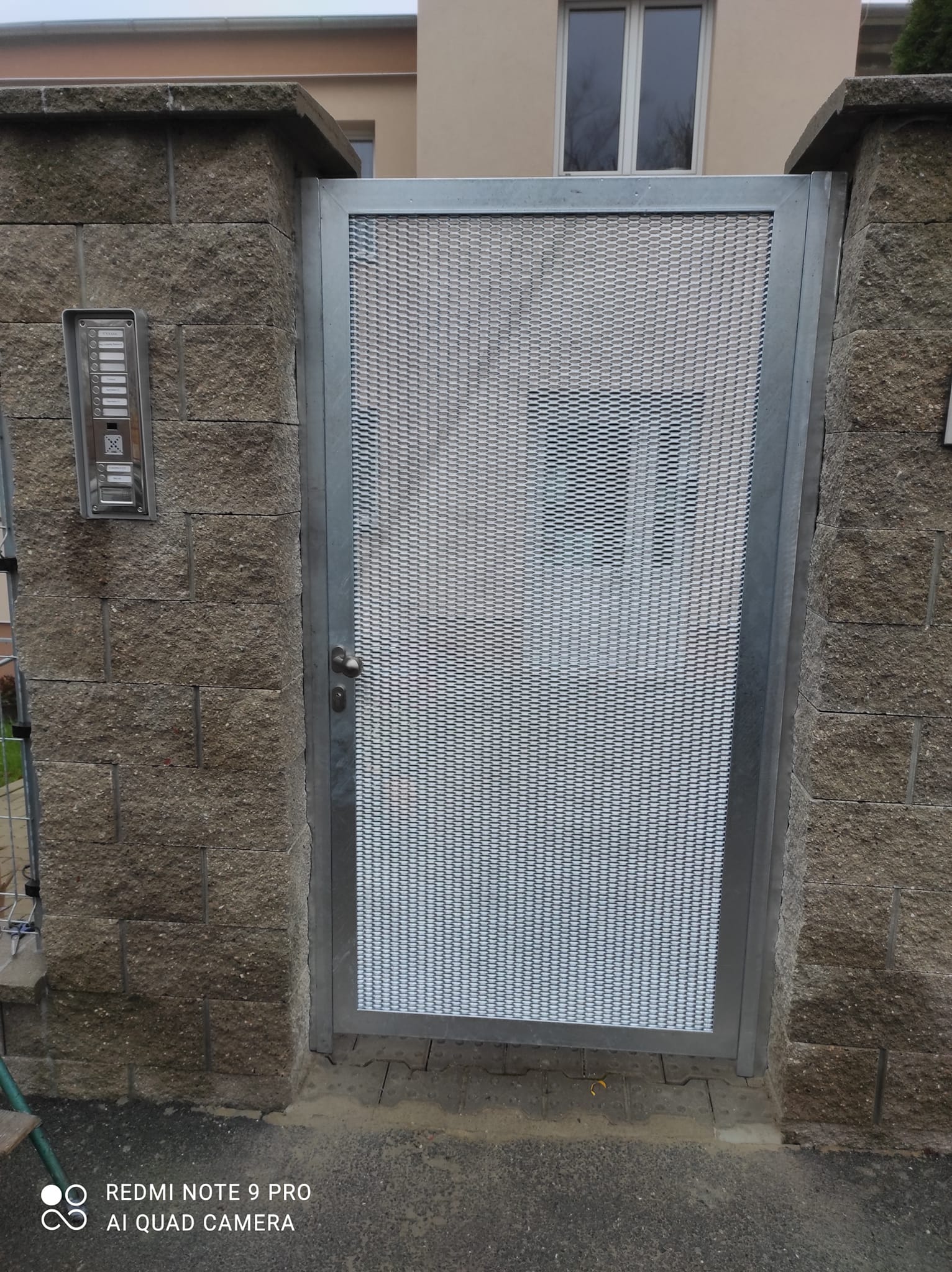 Čelní pohled na pozinkovanou branku mezi zděnými sloupky s panelem elektrického vrátného vedle vstupu.