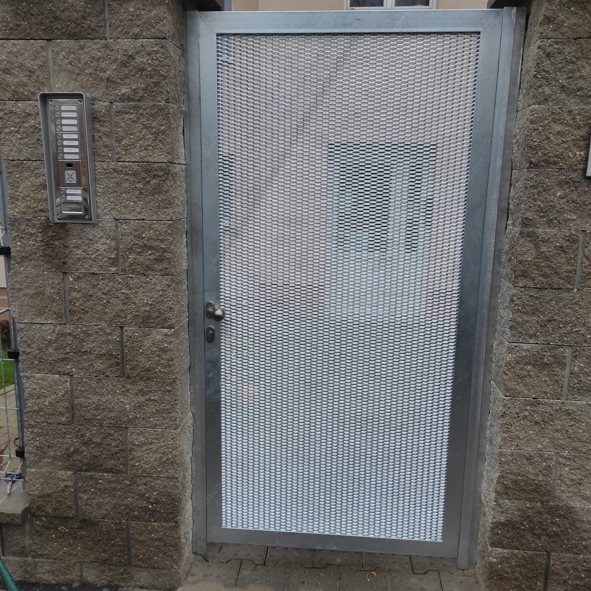 Čelní pohled na pozinkovanou branku mezi zděnými sloupky s panelem elektrického vrátného vedle vstupu.