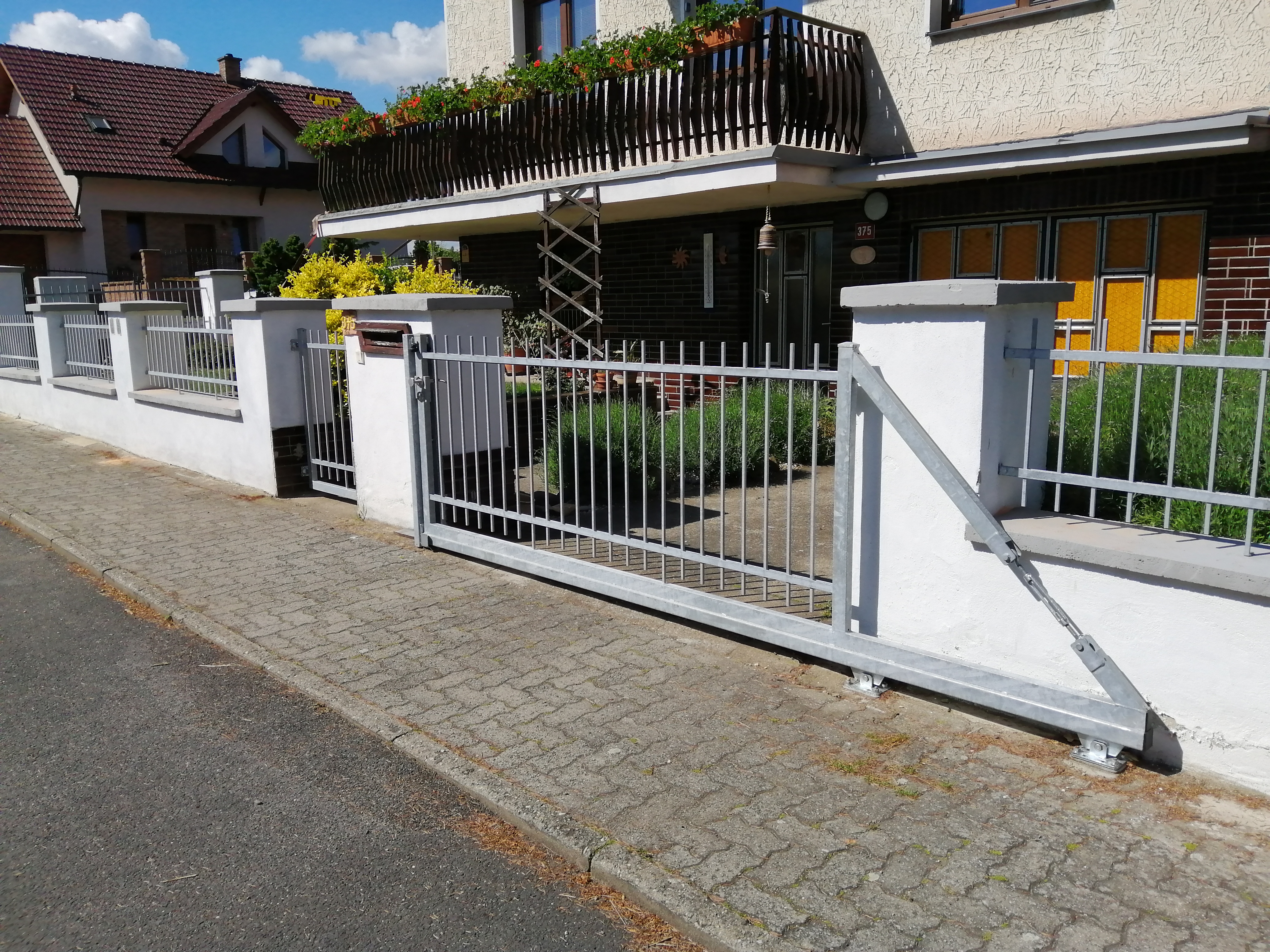 Detailní pohled z ulice na vjezdovou bránu, sousední branku a kovové výplně mezi zděnými sloupky.