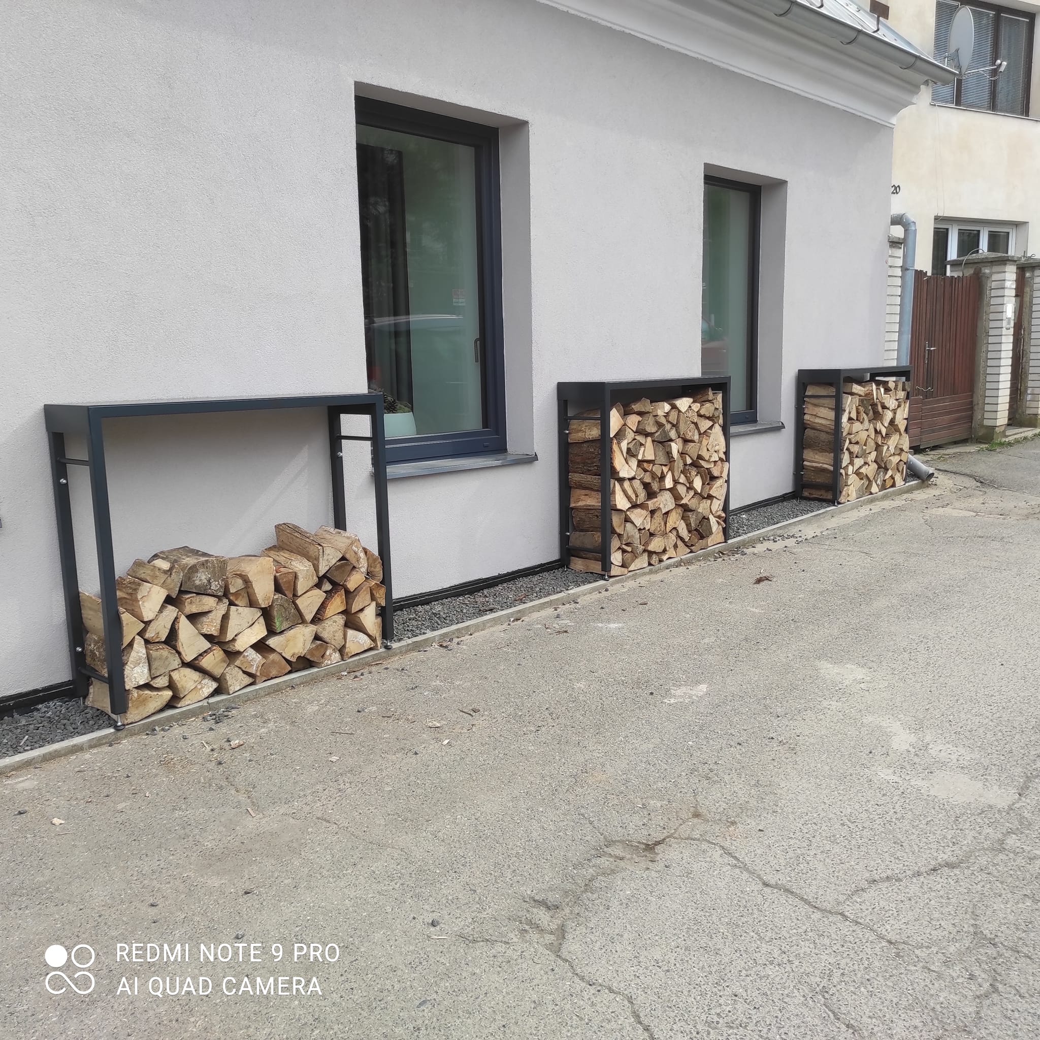 Tři kovové dřevníky mezi okny restaurace ve Statenicích, jeden částečně a dva více naplněné poleny.