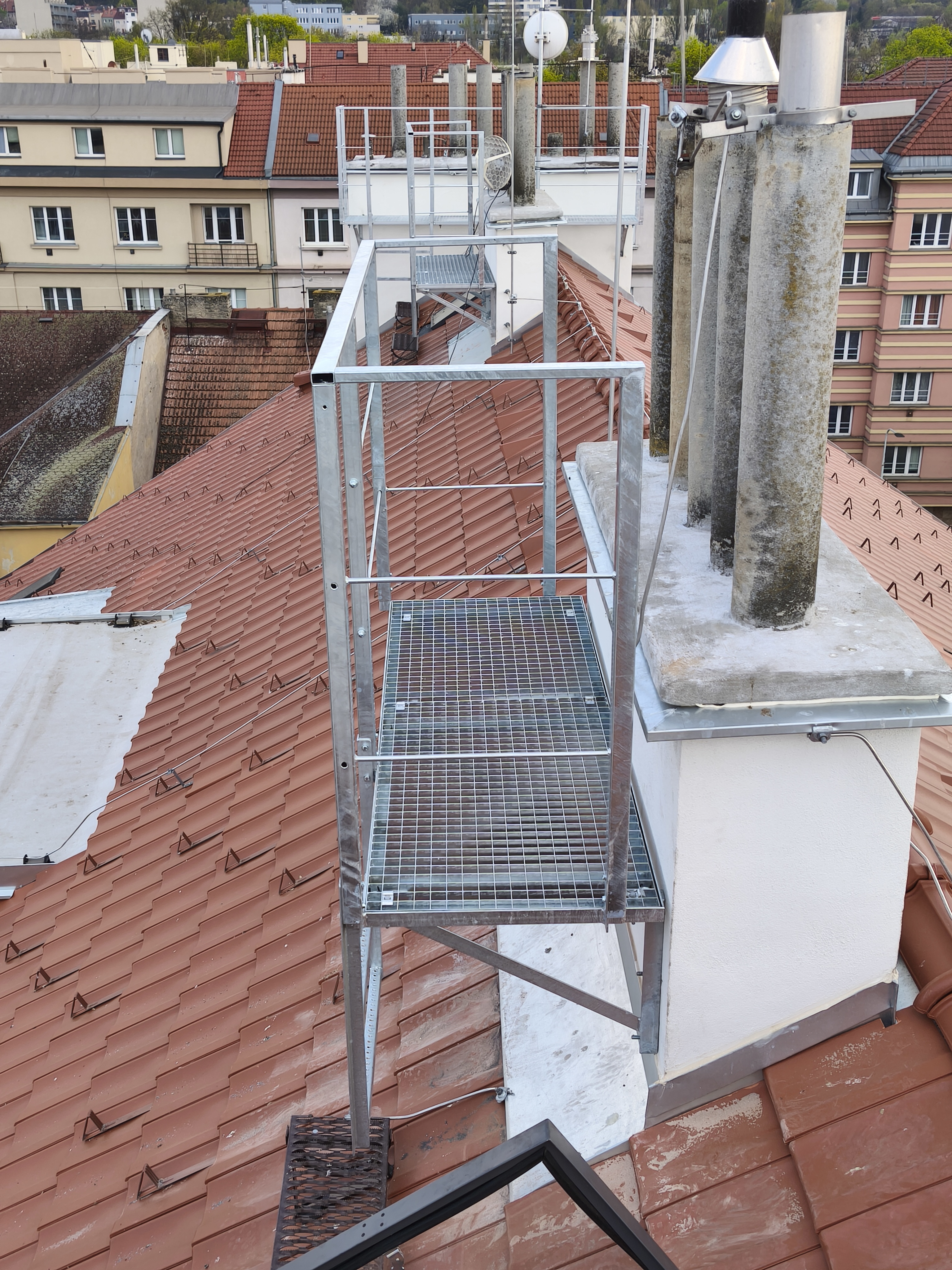 Komínové lávky s pororoštem a zábradlím rozmístěné u komínů nad šikmou střechou obytného domu.