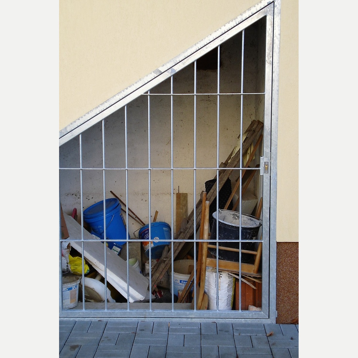 Žárově zinkovaná otevíravá mříž se šikmou horní hranou a svislou tyčovou výplní v úložném prostoru pod schodištěm ve Zlonicích.