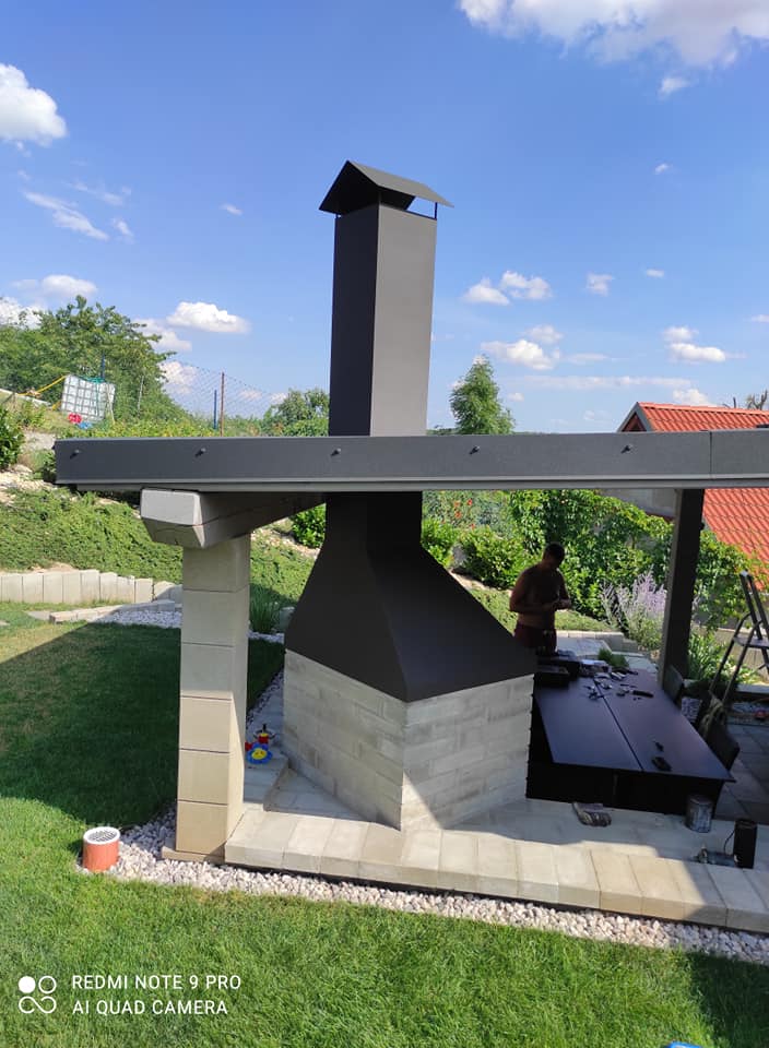 Černý kovový nástavec nad zděným venkovním krbem se svislým odvodem kouře při pohledu na celé posezení.