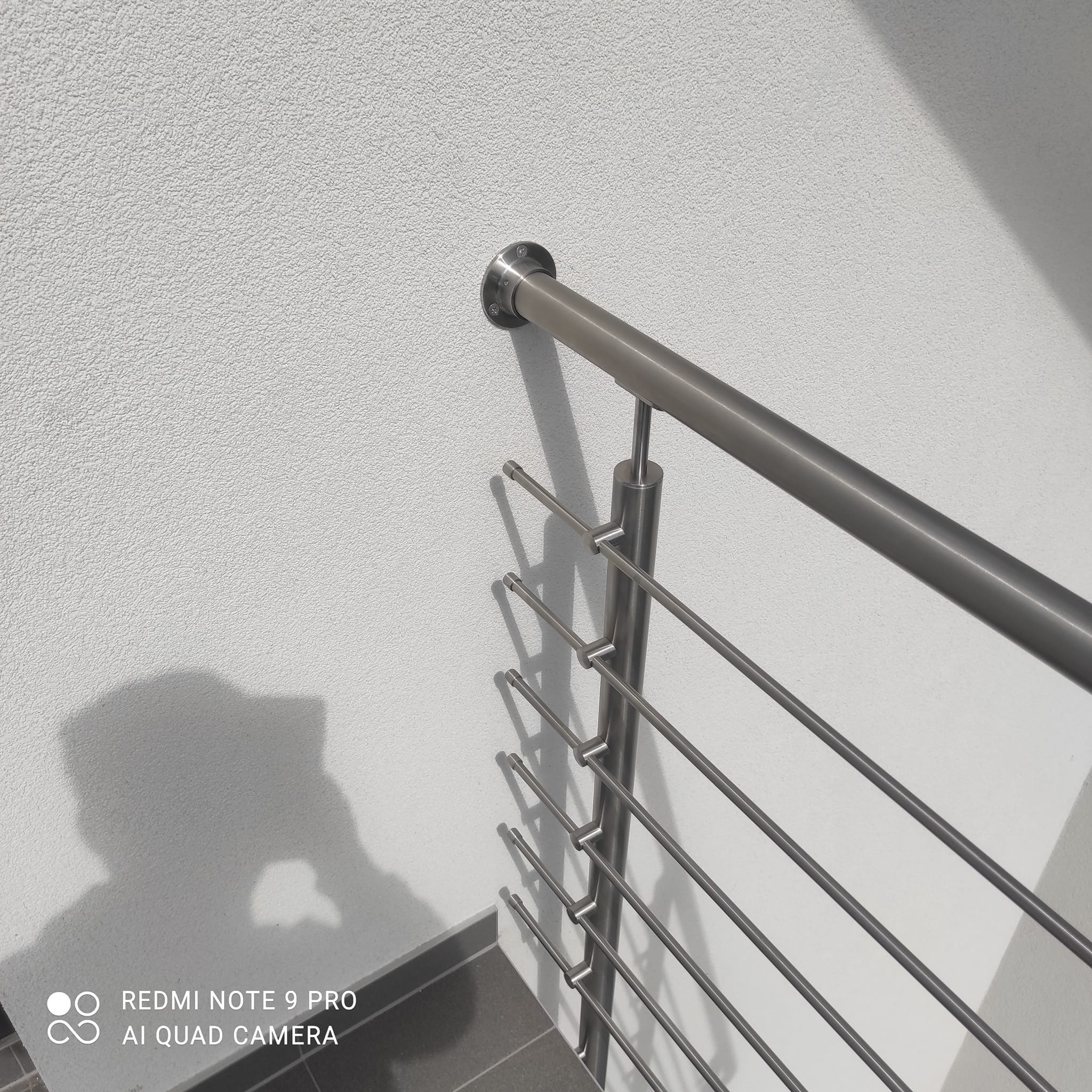 Detail ukončení nerezového madla u fasády a vodorovné tyčové výplně balkónového zábradlí.