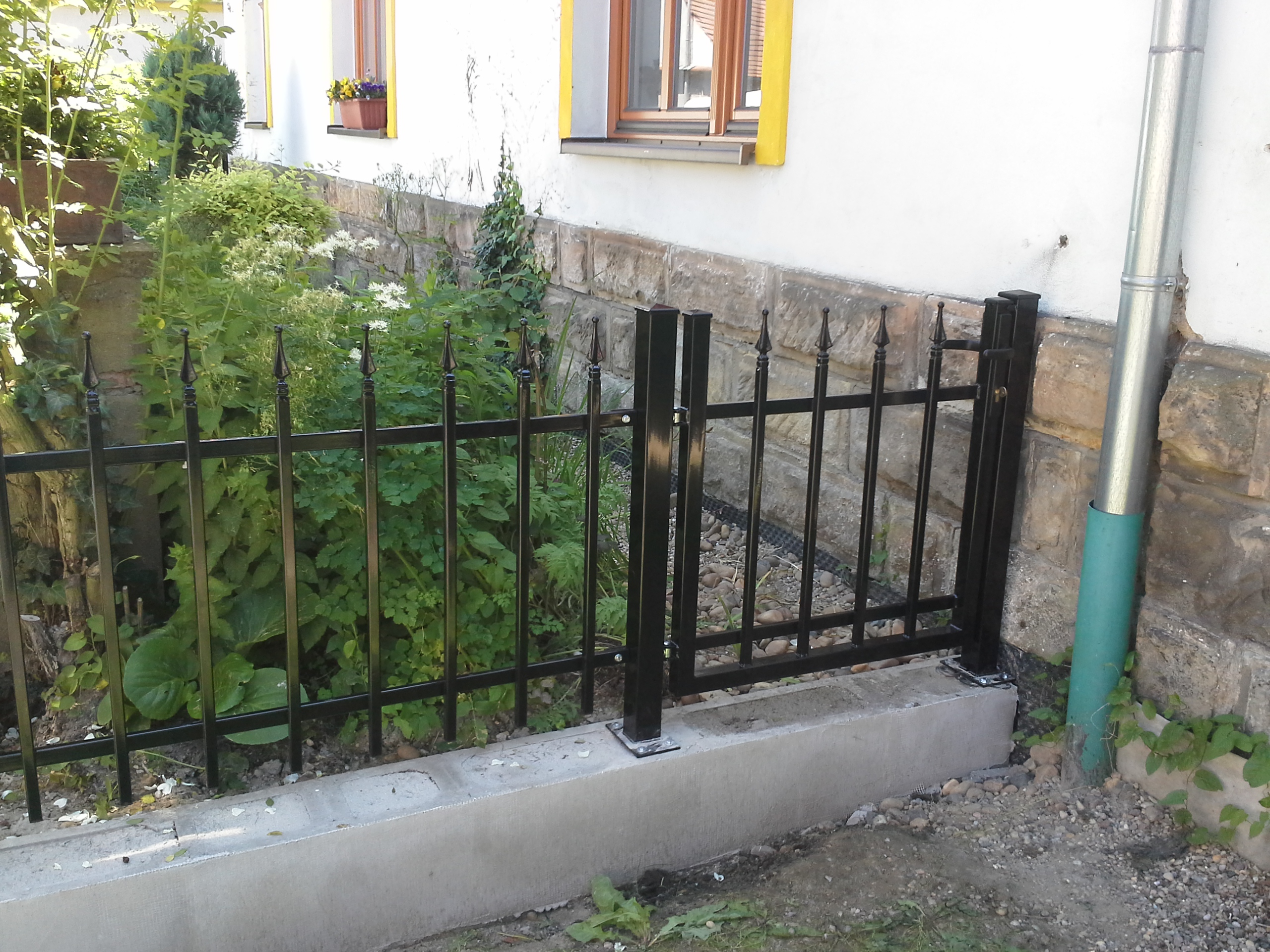Vstupní branka v černém oplocení předzahrádky, svislé profily zakončené ozdobnými špičkami.