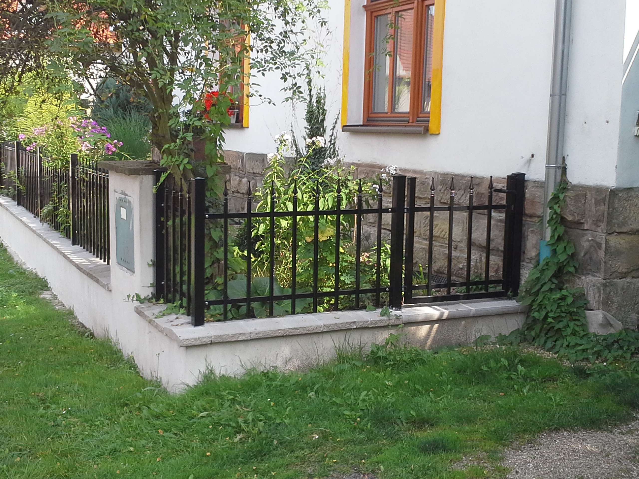 Rohové řešení černého kovového oplocení předzahrádky s malou vstupní brankou u stěny domu.