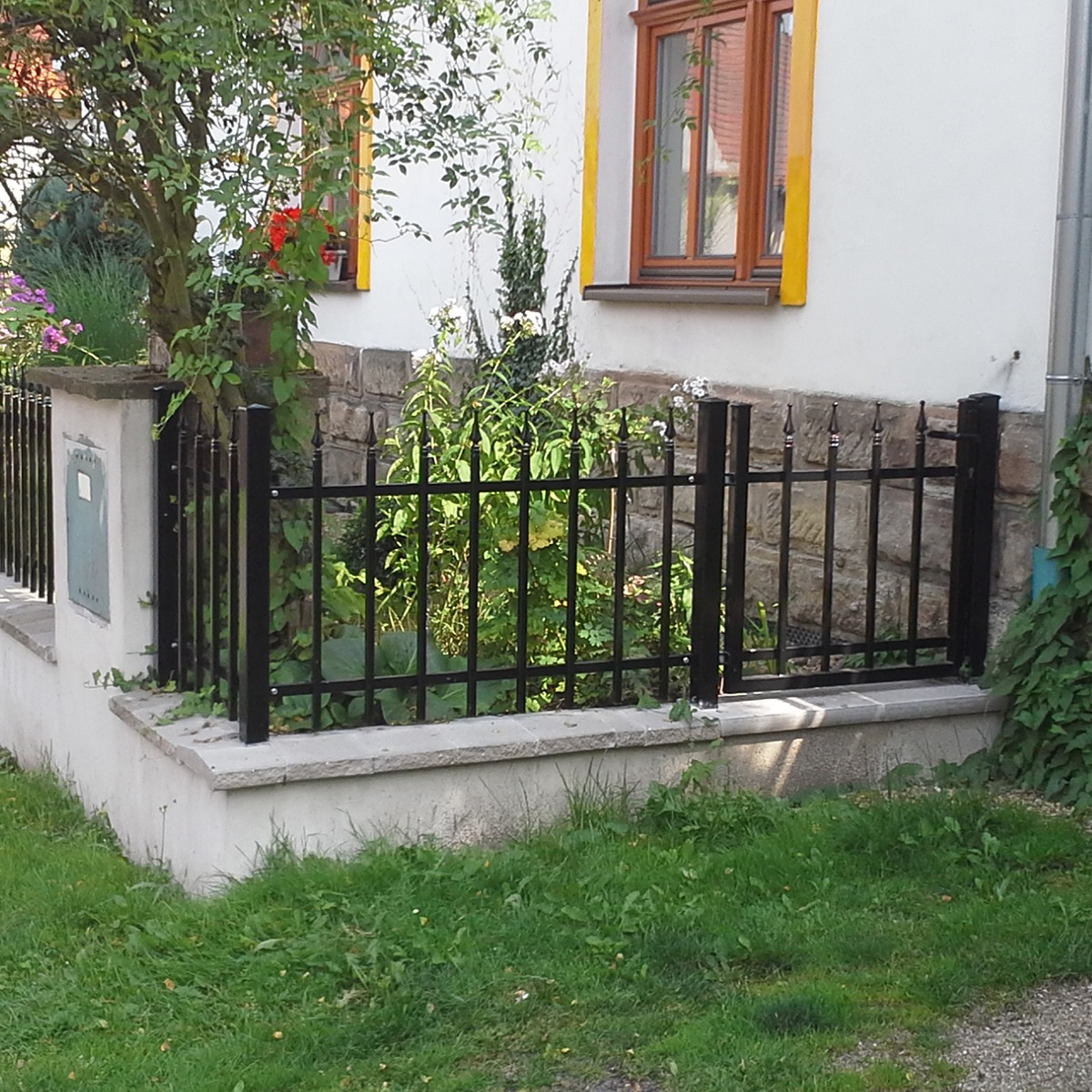Rohové řešení černého kovového oplocení předzahrádky s malou vstupní brankou u stěny domu.
