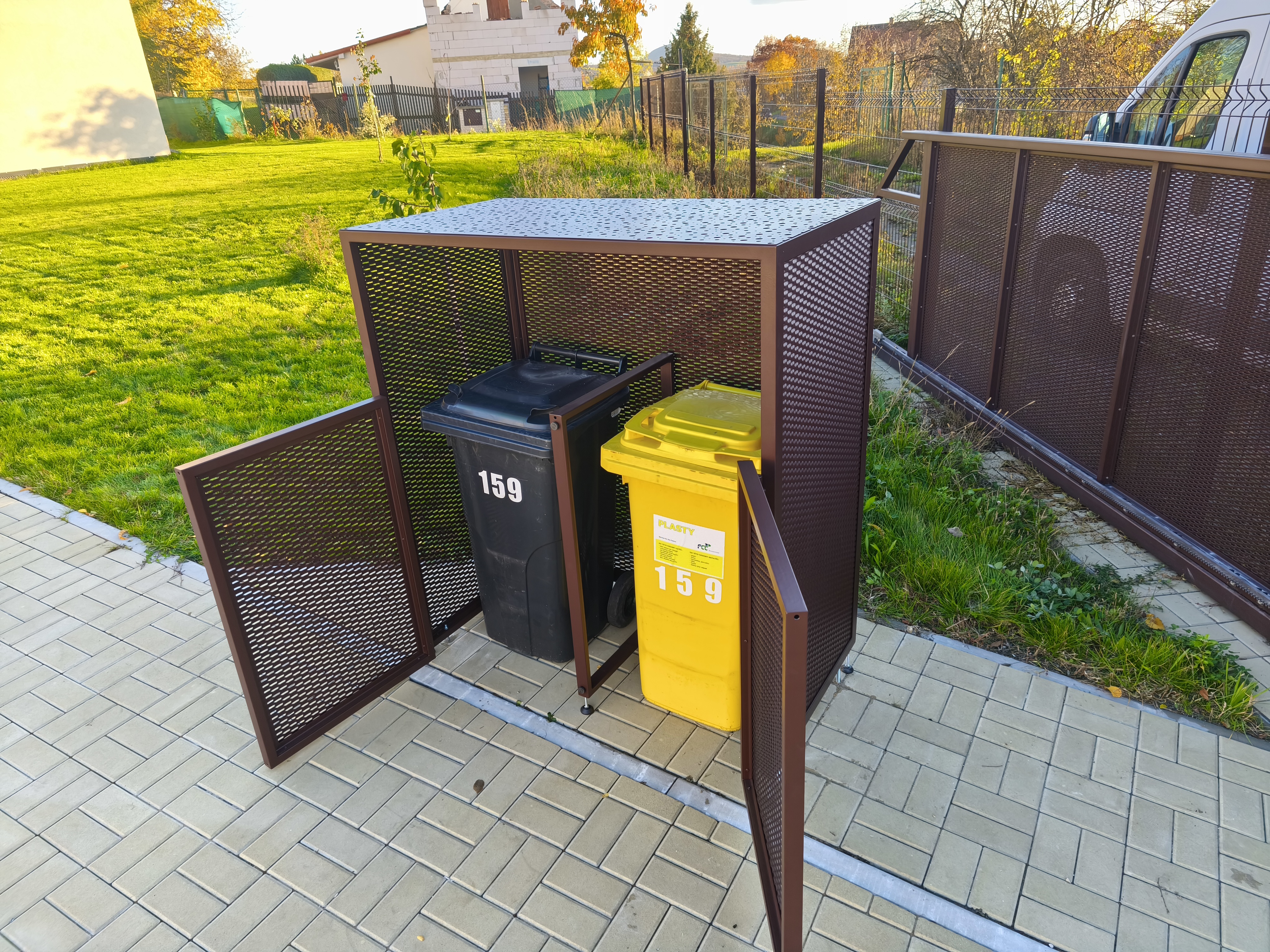 Otevřená čelní dvířka přístřešku a dvě popelnice uložené pod stříškou.