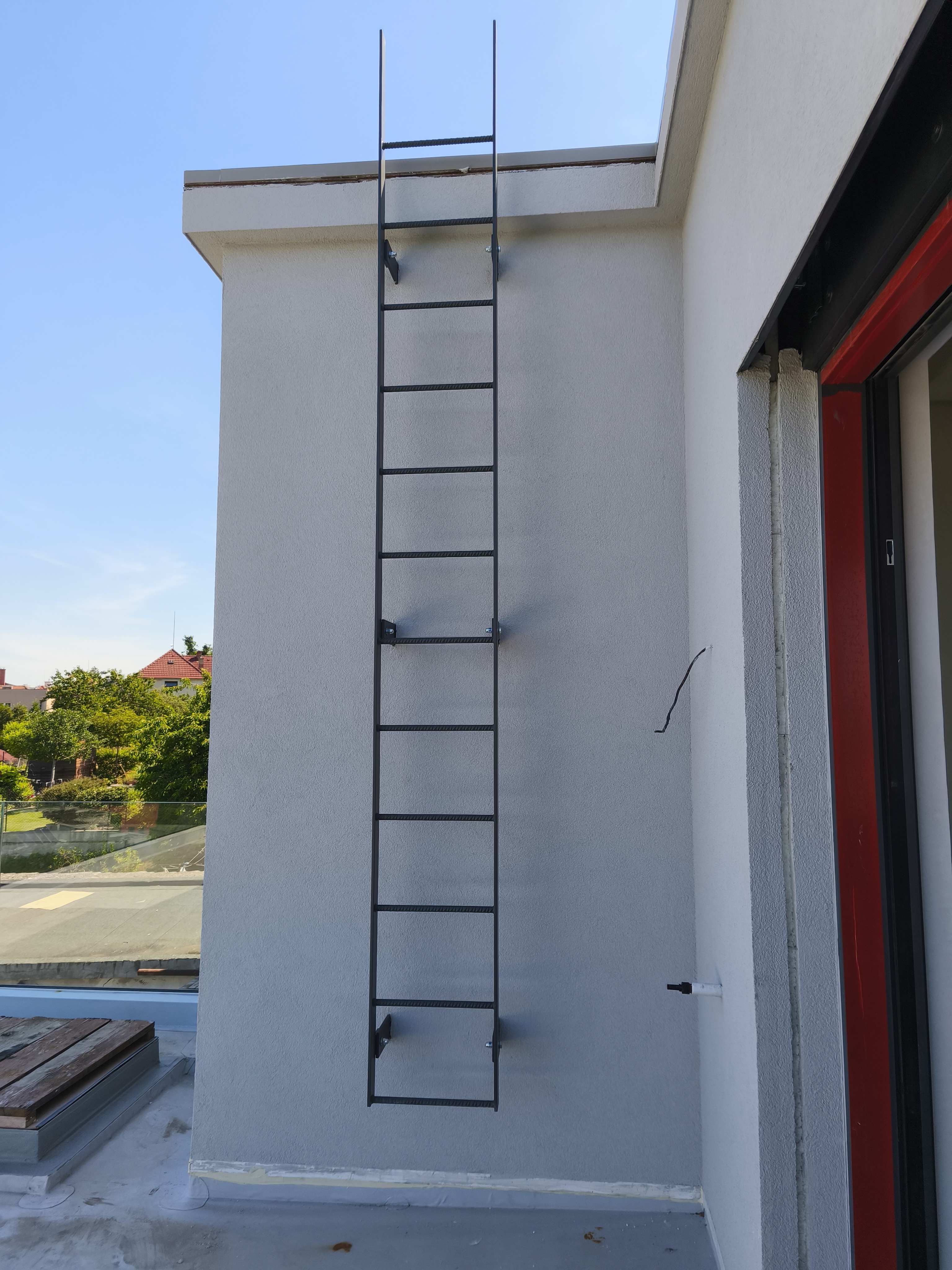 Antracitový servisní žebřík kotvený k fasádě s bočnicemi přesahujícími hranu střechy.