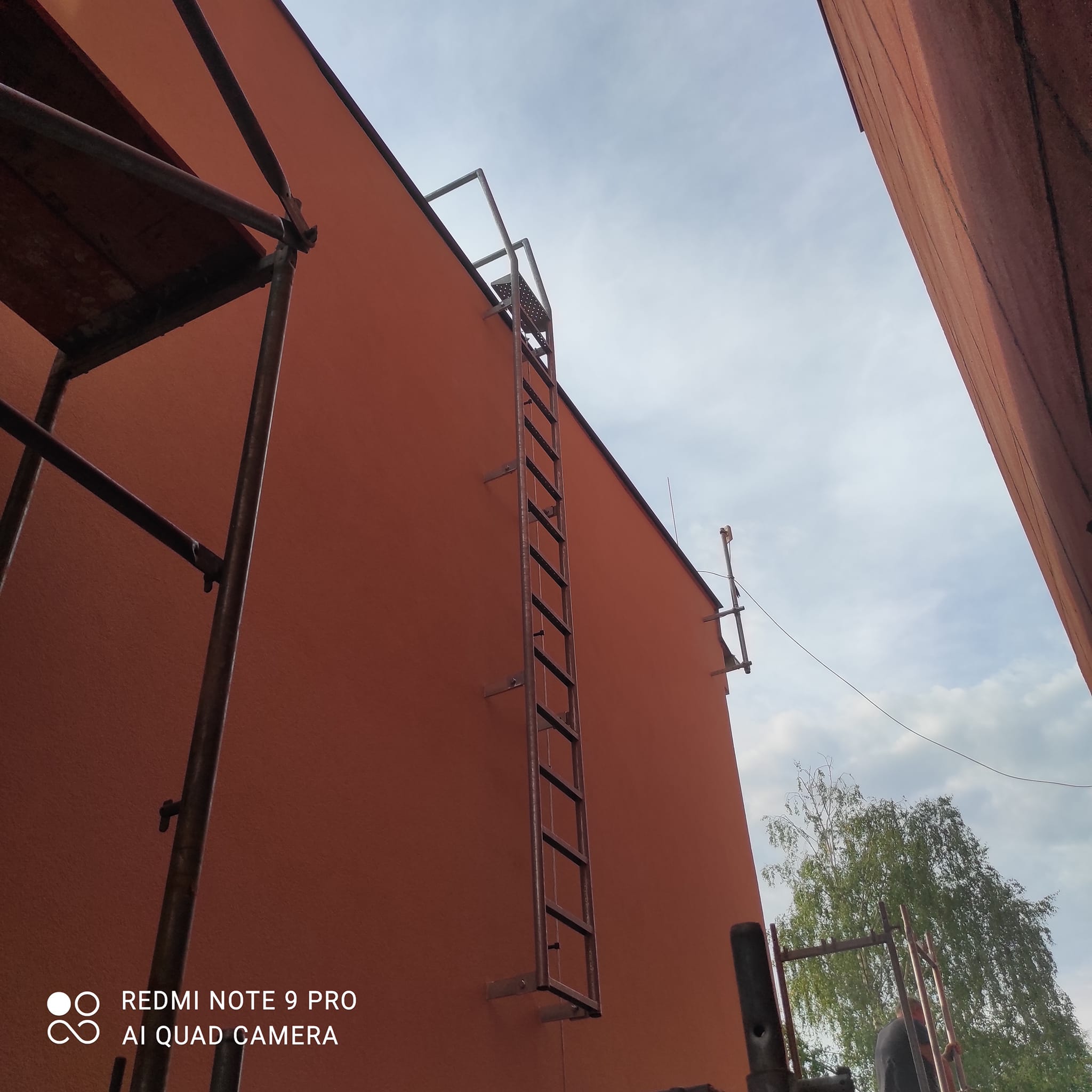 Celý servisní žebřík bez ochranného koše ukotvený u červené fasády a vedoucí na střechu.
