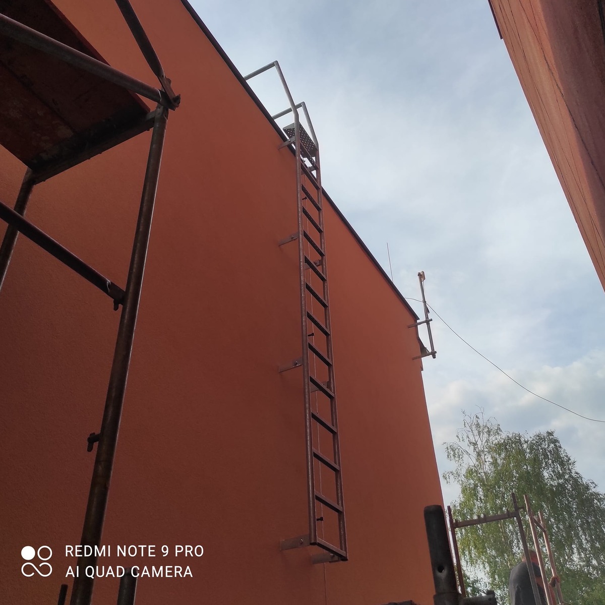 Celý servisní žebřík bez ochranného koše ukotvený u červené fasády a vedoucí na střechu.